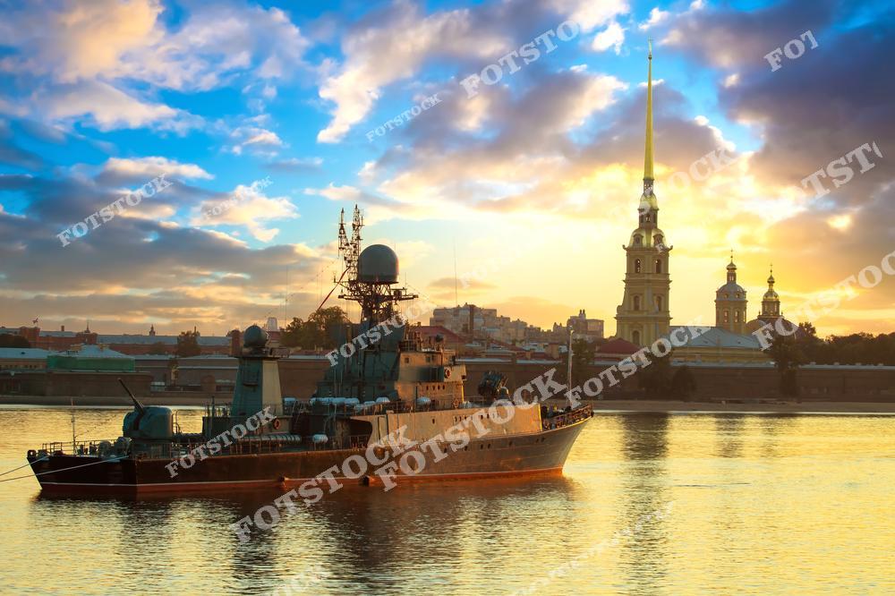 На фотографии изображен военный корабль, стоящий на Неве на фоне Петропавловской крепости. Золотой шпиль собора красиво выделяется на рассветном небе, наполненном облаками и теплым светом. Вода отражает яркие краски рассвета, создавая живописную композицию. Этот вид передает гармонию исторической архитектуры и морской традиции Санкт-Петербурга.